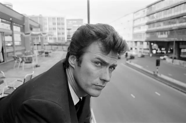 Una imagen en blanco y negro de Clint Eastwood asomado al balcón del Hotel Albany en Smallbrook Queensway, Birmingham, en junio de 1967, durante una gira promocional de "Por un Puñado de Dólares". Se pueden ver las sillas del balcón detrás de él, así como los edificios del Ringway Centre. Viste traje oscuro y camisa blanca.