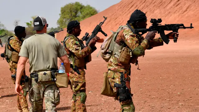 Un instructeur militaire américain passe devant des soldats maliens qui pointent leurs armes. Ils portent des tenues de camouflage et se tiennent devant une colline de terre rouge. La photo a été prise en 2018.