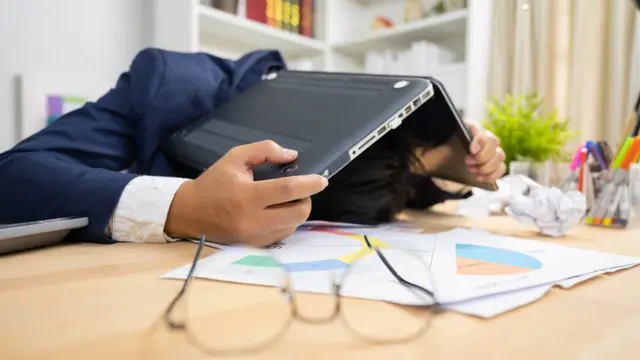 A man holds a computer over his head at a desk
