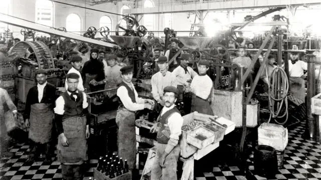 A imagem em preto e branco mostra o interior de uma fábrica no início do século 20, com dezenas de trabalhadores distribuídos entre grandes máquinas industriais. Muitos usam aventais e bonés, enquanto operam equipamentos ou posam para a fotografia. O ambiente é amplo, iluminado por janelas altas, e transmite o ritmo intenso da produção industrial da época.