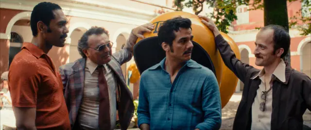 Quatro homens conversam ao ar livre, ao lado de uma cabine telefônica pública amarela. Um deles apoia a mão no topo da cabine enquanto fala com os outros. O homem ao centro, de bigode e camisa azul, olha para o lado com expressão atenta. Ao fundo, aparecem árvores e um prédio de arquitetura antiga com arcos, sugerindo uma praça ou pátio urbano.