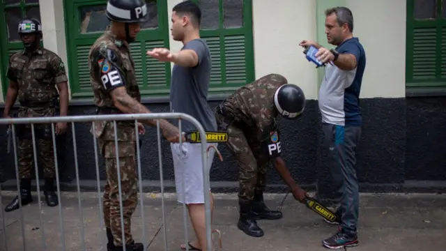 Policiais passam detectoresmetais perto dos corposdois homens, do ladoforauma construção