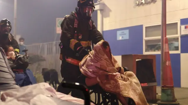 車いすに座り毛布にくるまった患者が、屋外で消防隊員に車いすを押されて避難している。近くには別の患者らしき人や、黒い犬などがいる