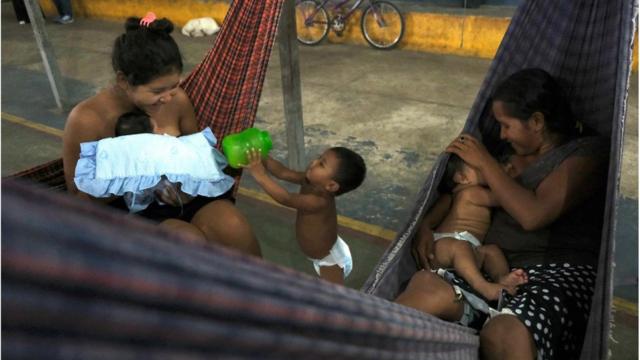 Duas mulheres warao, sentadas em redes, com bebês no colo e uma criança de frauda entre as redes