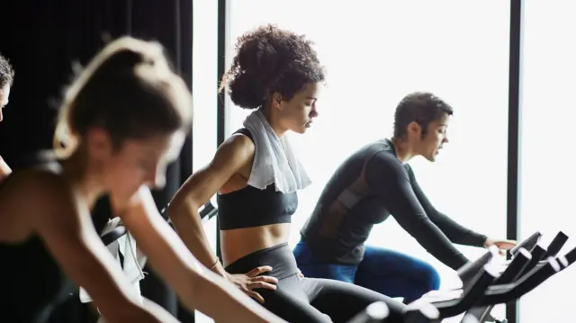 Pipo wey dey do indoor exercise bike. Di young woman for middle dey rest, her hand dey for her hips. She get towel for her neck.