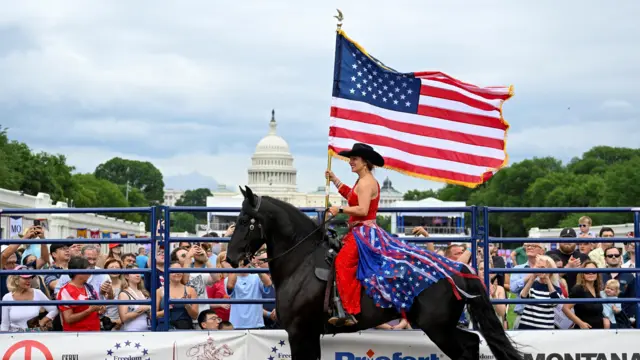 elinde amerikan bayrağı tutan bir kadın at üzerinde renkli rodeo kıyafetiyle, etrafında insanlar onu izliyor ve arkada kongre binası görülüyor