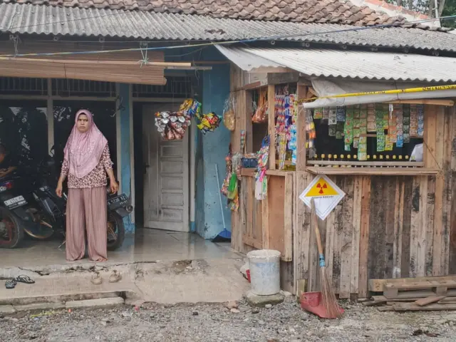 Seorang perempuan berdiri di depan rumahnya. Di warung kecilnya terdapat stiker bahaya radiasi.