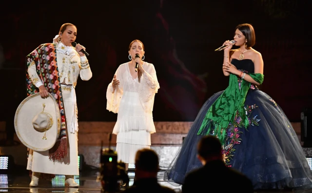 A la izquierda, Aida Cuevas, vestida con una traje de mariachi blanco, con un sombrero en la mano y en la otra, un micrófono mientras canta. En el centro está Natalia Lafourcade, con un vestido blanco, también canta con un micrófono en la mano, y a la derecha, Ángela Aguilar, con un vestido negro adornado con flores de colores y un chal verde