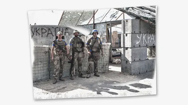 Трое мужчин в военной форме с оружием стоят у бетонного заграждения, на котором написано краской "Украiна"