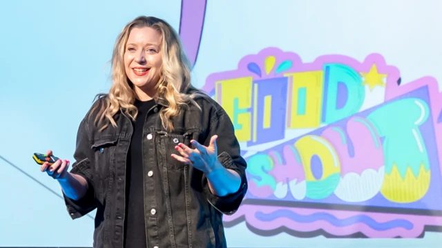 ኤሚ ኪን በቀለማት ካሸበረቀ ስክሪን ፊት ለፊት ቆማ ታዳሚዎች ንግግር እያደረገች
