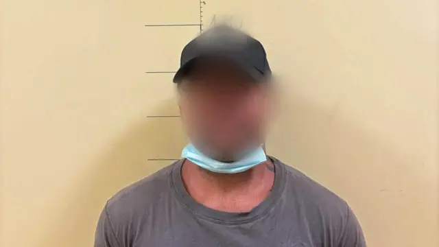 Mężczyzna w czarnej czapce z daszkiem i ciemnym t-shircie stoi na tle żółtej ściany. Jego twarz jest zamazana, a pod brodą ma założoną maseczkę chirurgiczną.