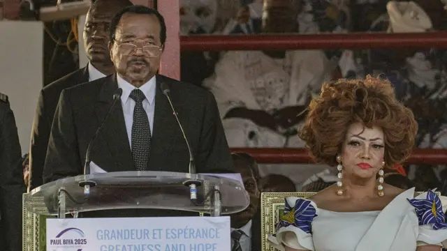President Paul Biya stands at a lectern and speaks into a pair of microphones. He is flanked by his wife, Chantal Biya. 