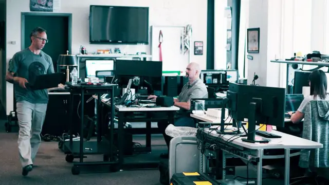 গ্রেগ স্কয়ার এবং পিট ম্যানিং-সহ তাদের অফিসের সহকর্মীরা। গ্রেগ একটি সবুজ টি-শার্ট এবং হালকা প্যান্ট পরে আছেন এবং একটি ল্যাপটপ ধরে কম্পিউটার স্ক্রিনে ভরা একটি ঘরের মধ্য দিয়ে হেঁটে যাচ্ছেন।