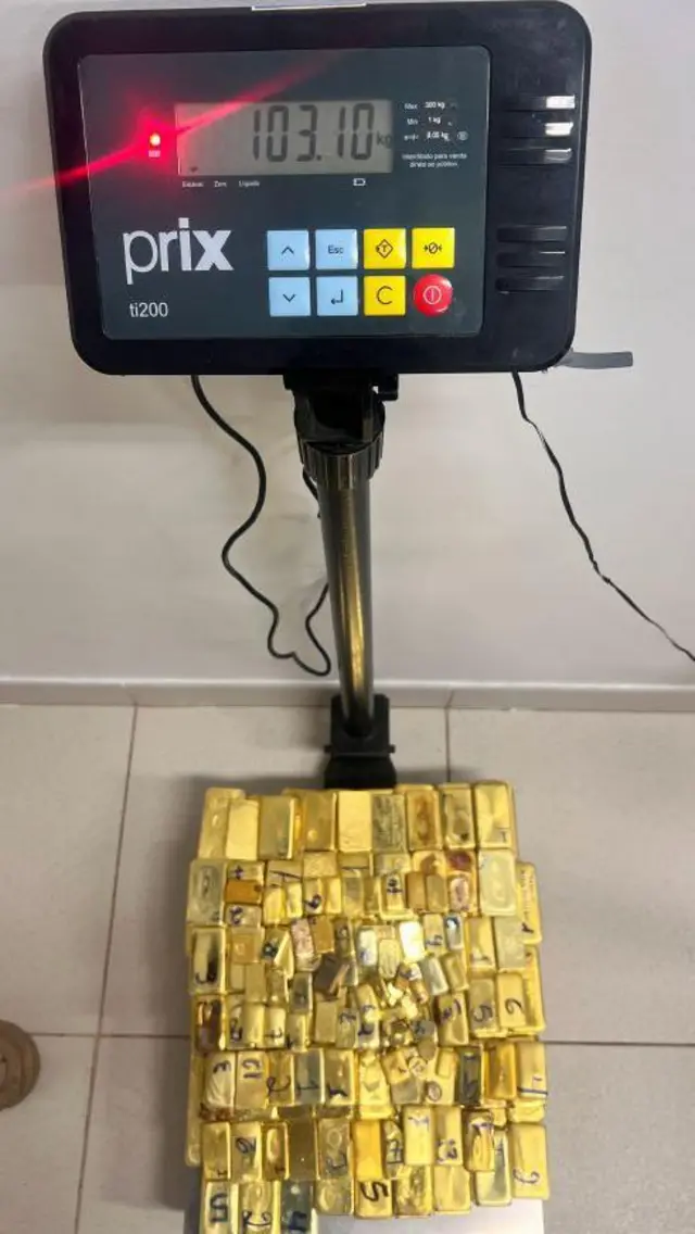 Balança marcando 103,1 quilos em barras de ouro diante de uma parede branca