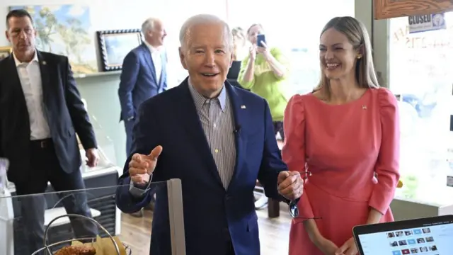 На переднем плане корзинка с едой и компьютер, в центре композиции улыбающийся Джо Байден, справа стоит смеющаяся Когнетти в красном платье, на заднем фоне видны другие люди.