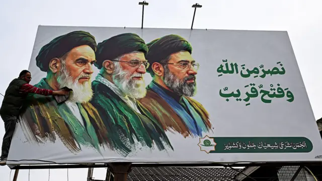 İran’ın hayatını kaybeden dini liderleri Ayetullah Ruhullah Humeyni (solda) ve Ayetullah Ali Hamaney’in (ortada) yer aldığı, yeni seçilen dini lider Ayetullah Mücteba Hamaney’in de bulunduğu bir reklam panosunu temizleyen bir adam.