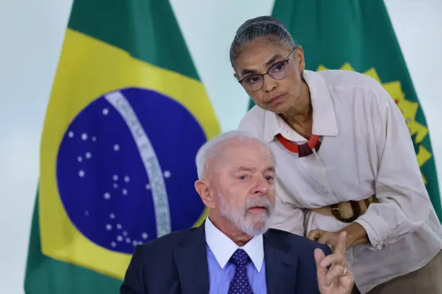 Lula e Marina Silva próximos, falando entre si. Ela está em pé, agachando em direção a ele, que está sentado