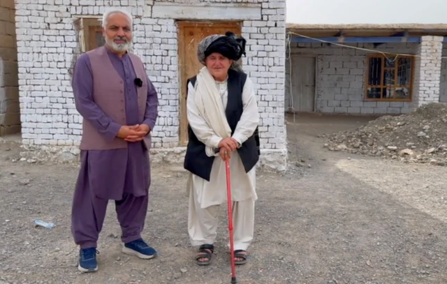 BBC мухбири эркак кийимини кийиб салла ўраган, ҳаса ушлаган, 70 ёшли Ҳукмина билан, Ҳукминанинг уйи олдида туришибди