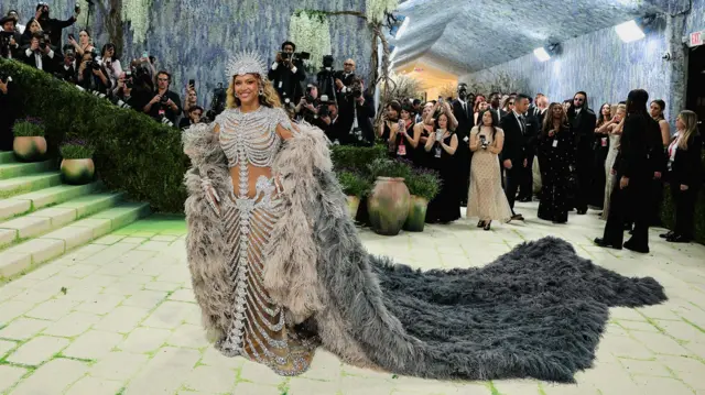 Beyoncé ma na sobie bogato zdobioną, jasną suknię z motywem szkieletu oraz długą, obszerną pelerynę z szarego futra, ciągnącą się po ziemi. W tle widać schody i ściany udekorowane zielenią oraz wiszącymi roślinami, a także tłum fotoreporterów i gości obserwujących wydarzenie.