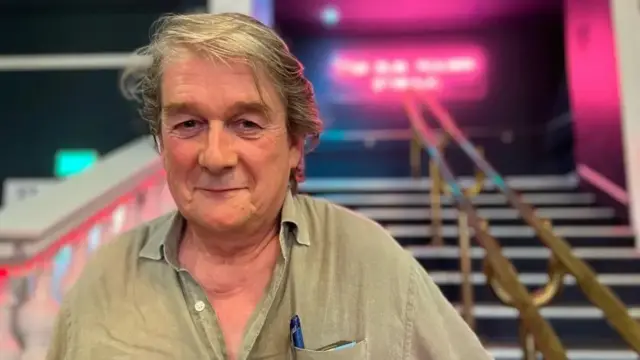 Hugh se encuentra frente a una escalera con pasamanos a ambos lados, que conduce a una zona iluminada con luces rosas y moradas. Lleva una camisa abotonada de color claro y un bolígrafo en el bolsillo. Se ve un texto de neón al fondo, pero está parcialmente oculto por los efectos de iluminación.

