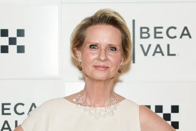 Cynthia Nixon stands in front of a sign wearing a white gown