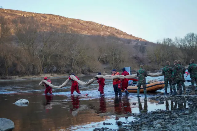 Група рятувальників і людей у камуфляжі стоїть у неглибокій воді річки. Вони несуть довгий гнучкий бар’єр, імовірно для стримування забруднення. На березі поруч видно надувний човен. У тлі – пагорб і дерева без листя.