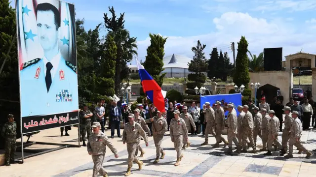 Russian soldiers parade in front of a poster of Syrian president Bashar al-Asad during a ceremony marking Soviet victory over Nazi Germany in WW II, in Aleppo's Saadallah al-Jabiri square in northern Syria on May 9, 2018.