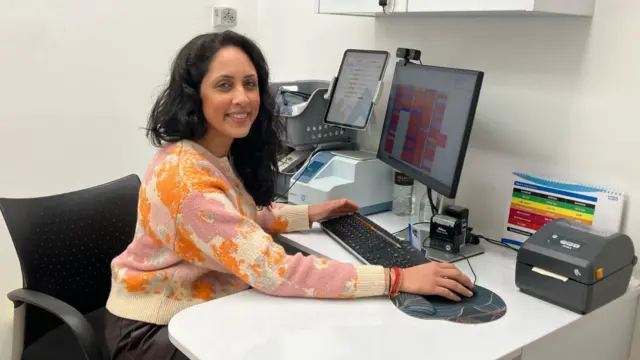 Hema Patel está sentada en un escritorio blanco sobre una silla de oficina negra. Tiene un monitor, un teclado y un ratón delante, y apoya la mano en el ratón. También hay otros dispositivos periféricos sobre el escritorio. Lleva un jersey claro con estampados rosas y naranjas. Tiene el pelo negro y sonríe a la cámara.