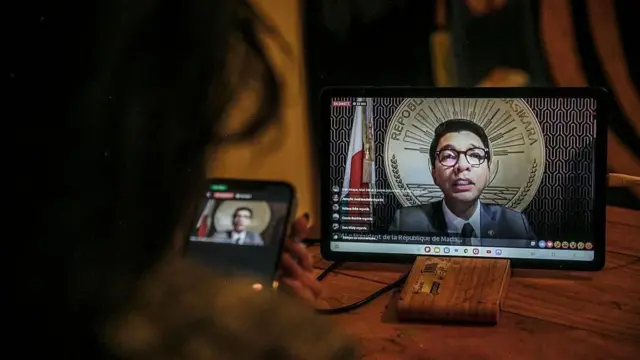 机に置かれたタブレットの画面に、ラジョエリナ大統領の演説配信が映っている。机の前に爪の長い人物が座り、左手で持ったスマートフォンで同じ演説の映像を見ているのが、真後ろから撮られている