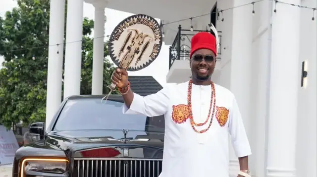 Foto Obi Cubana dịka o ji akupe n'aka, yiri akwa ọcha ma na-achị ọchị n'ihu ụgbọala ya