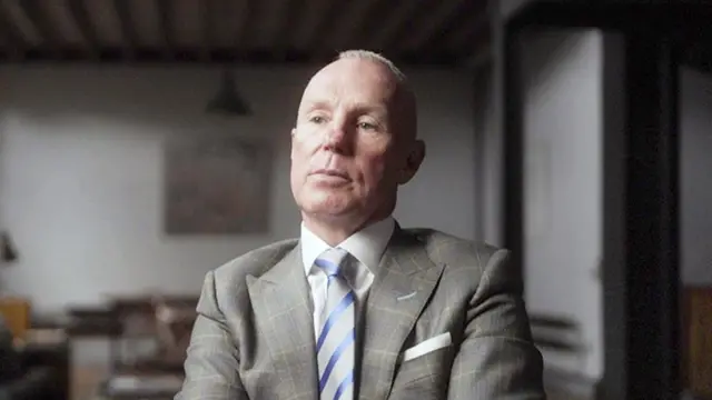 罗恩·布莱克身穿浅灰色格纹西装,搭配白色衬衫与蓝色条纹领带,坐在室内一间光线柔和的房间内,天花板有木质横梁,背景中可见模糊的家具。