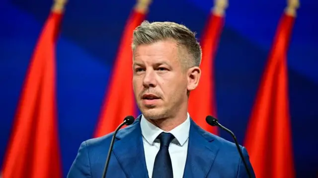 Čovek u plavom odelu ispred zastava i plave pozadine