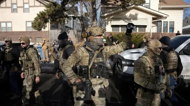 一軒家が並ぶ住宅街に、重装備で顔を隠した迷彩服姿の当局者が複数立っている
