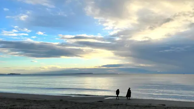 Se ven las siluetas de dos personas caminando por una playa a la sombra del sol, oculta tras unas nubes ligeras. El agua está tranquila, con apenas algunas olas en la superficie.
