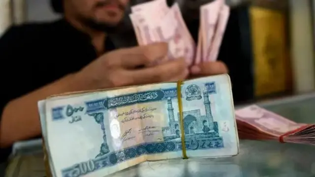 د افغانیو د زرګون او پنځه سوه ګون بنډل په مېز ایښی او بک‌ګروند کې یو سړی چې ظاهرآ صراف دی، پیسې شمېرې
