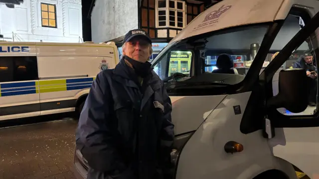 Un hombre canoso lleva una gorra azul marino con la inscripción "Pastores de la calle Stafford" escrita en blanco. Lleva una chaqueta azul marino a juego con la misma inscripción en el pecho. Está apoyado en una furgoneta blanca.