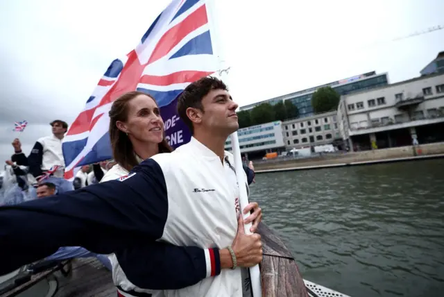 Les porte-drapeaux Tom Daley et Helen Glover, de l'équipe britannique, font un geste sur un bateau tout en tenant le drapeau national sur la Seine pendant la cérémonie d'ouverture.