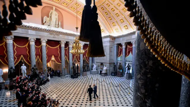 Широк кадар приказује краља како га воде кроз ходник Капитола, док су репортери са стране.