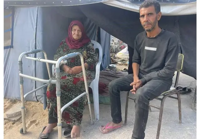 Rami y su madre sentados a la entrada de una carpa. La madre tiene junto a ella un andador para caminar.