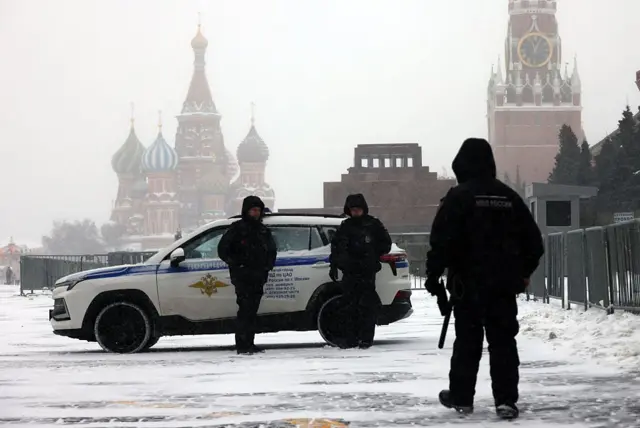 Засніжена Красна площа в Москві. На передньому плані – троє озброєних поліцейських поруч із білим позашляховиком. У глибині кадру видно собор Василія Блаженного, мавзолей Леніна та вежу Кремля з годинником. Погода похмура, мжичка зі снігом, огорожі обмежують рух перехожих.