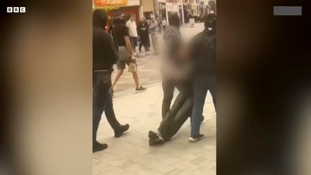 لقطة من مقطع فيديو توضح أفراداً في ملابس مدنية يرتدون أقنعة وهم يقتادون مشتبهاً به،و على ذراعيه أكياس بلاستيكية شفافة، عبر منطقة للمشاة