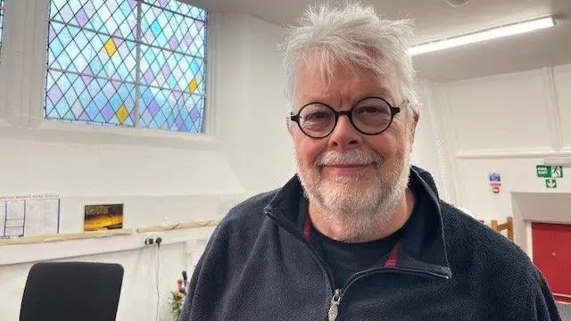 Un hombre con gafas redondas de montura negra sonríe a la cámara. Lleva una sudadera azul de forro polar con cremallera, y a su lado hay una vidriera.