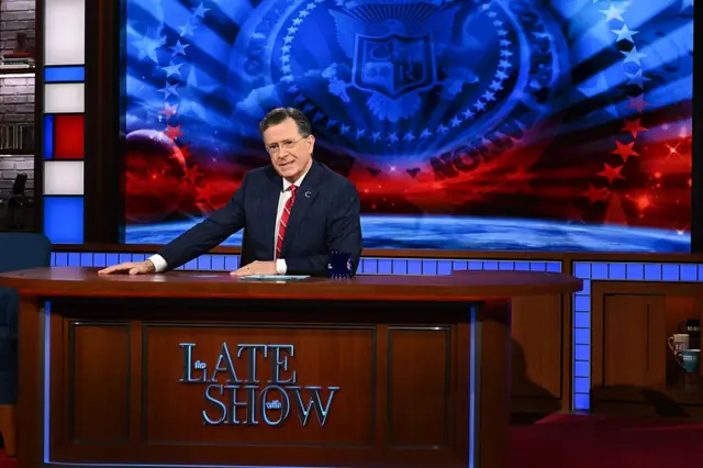 Stephen Colbert sentado detrás del escritorio de “Late Show”, con un fondo rojo y azul con estrellas.