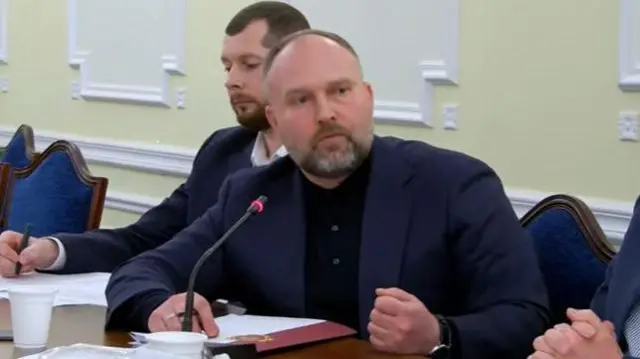 На передньому плані Євген Пронін, голова Держфінмоніторингу. Він дивиться праворуч за співрозмовника, що лишився за кадром. Пронін вдягнений у темно-синій діловий піджак та чорну сорочку. Ліва рука стиснена в кулак. 