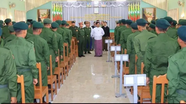 ဟောခန်းကျယ်ထဲမှာ ခုံတန်းအလိုက်တန်းစီမတ်တပ်ရပ်နေတဲ့ စစ်ဝတ်စုံနဲ့တပ်သားတွေ၊ တပ်သားတွေရဲ့ထိပ်မှာရပ်နေတဲ့ တိုက်ပုံနဲ့ပုဆိုးဝတ်အမျိုးသားနဲ့ စစ်ဝတ်စုံဝတ် အရာရှိတွေ