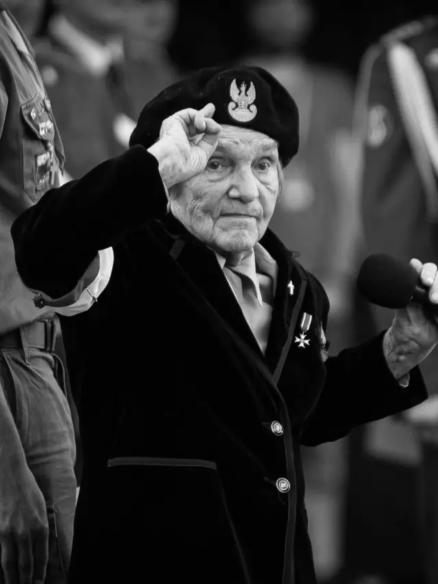 Чорно-біле фото літньої жінки у береті з польським орлом та нагородами на піджаку. Людина стоїть біля мікрофона й підносить руку до головного убору, віддаючи честь.