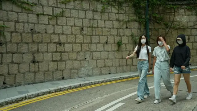Üç kadın, yüzlerinde maske takarak sakin bir sokakta yan yana yürüyor. Asfalt yol hafifçe kıvrılıyor, kenarları çift sarı çizgiyle, şeritleri ayıran beyaz bir çizgiyle belirlenmiş. Kadınlar açık renkli bluzlar, kot pantolonlar ve spor ayakkabılar gibi günlük kıyafetler giymişler.