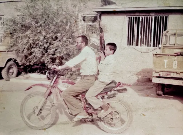 رضا شکرالهی در حالی که یک اسلحه کلاشینکف در دست دارد پشت موتورسیکلت همراه پدرش دیده می‌شود