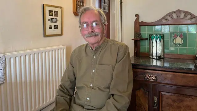 Мужчина с короткими седыми волосами и густыми седыми усами в очках в тонкой оправе одет в зеленую рубашку с длинными рукавами и сидит и смотрит в камеру. Позади него стоит деревянный винтажный шкаф, а слева — батарея и картины на стенах.