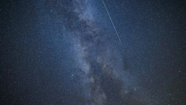 Perseid meteor yağmurunun zirve noktasında, koyu mavi gece gökyüzünün ortasında, karakteristik yeşil izini bırakan beyaz bir meteor, Samanyolu'nun önünde görülüyor.